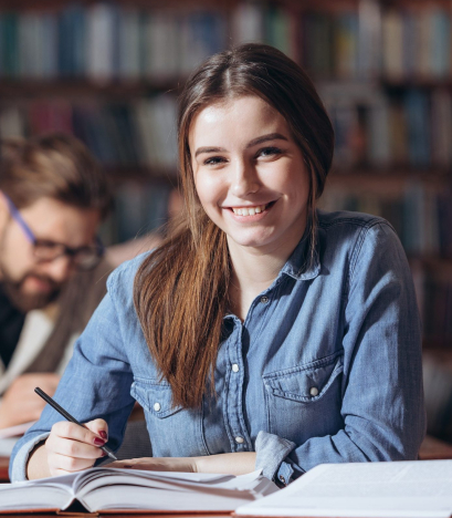 Attractive girl with long brown hair in jeans shirt sitting at desk and taking notes from open books. Female student smiling and looking at camera in public library.